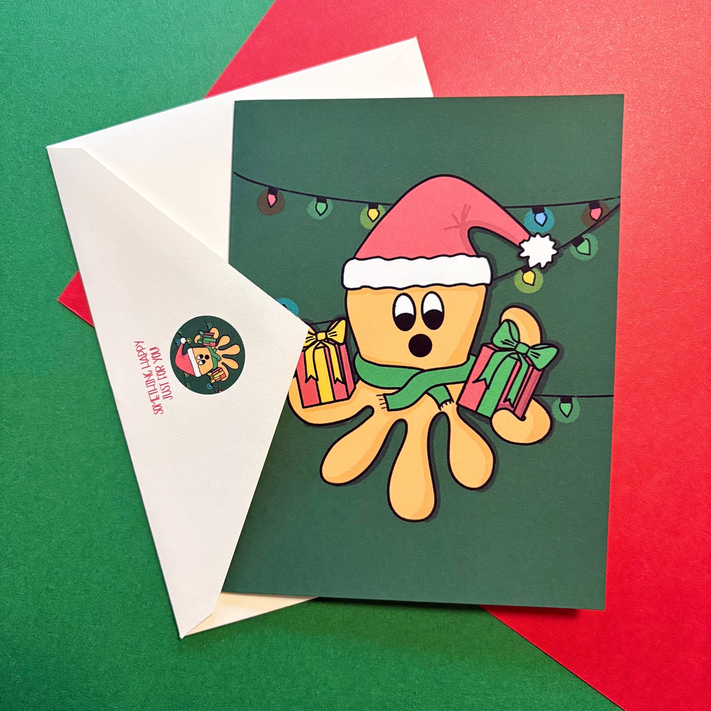 Christmas-themed card and envelope featuring an illustrated orange octopus wearing a santa hat and holding presents on a green background