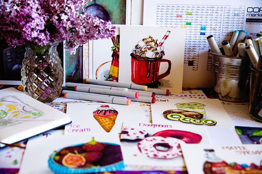 an artist's desktop with several in progress food illustrations, some copic markers and swatch charts and a vase of lilac flowers