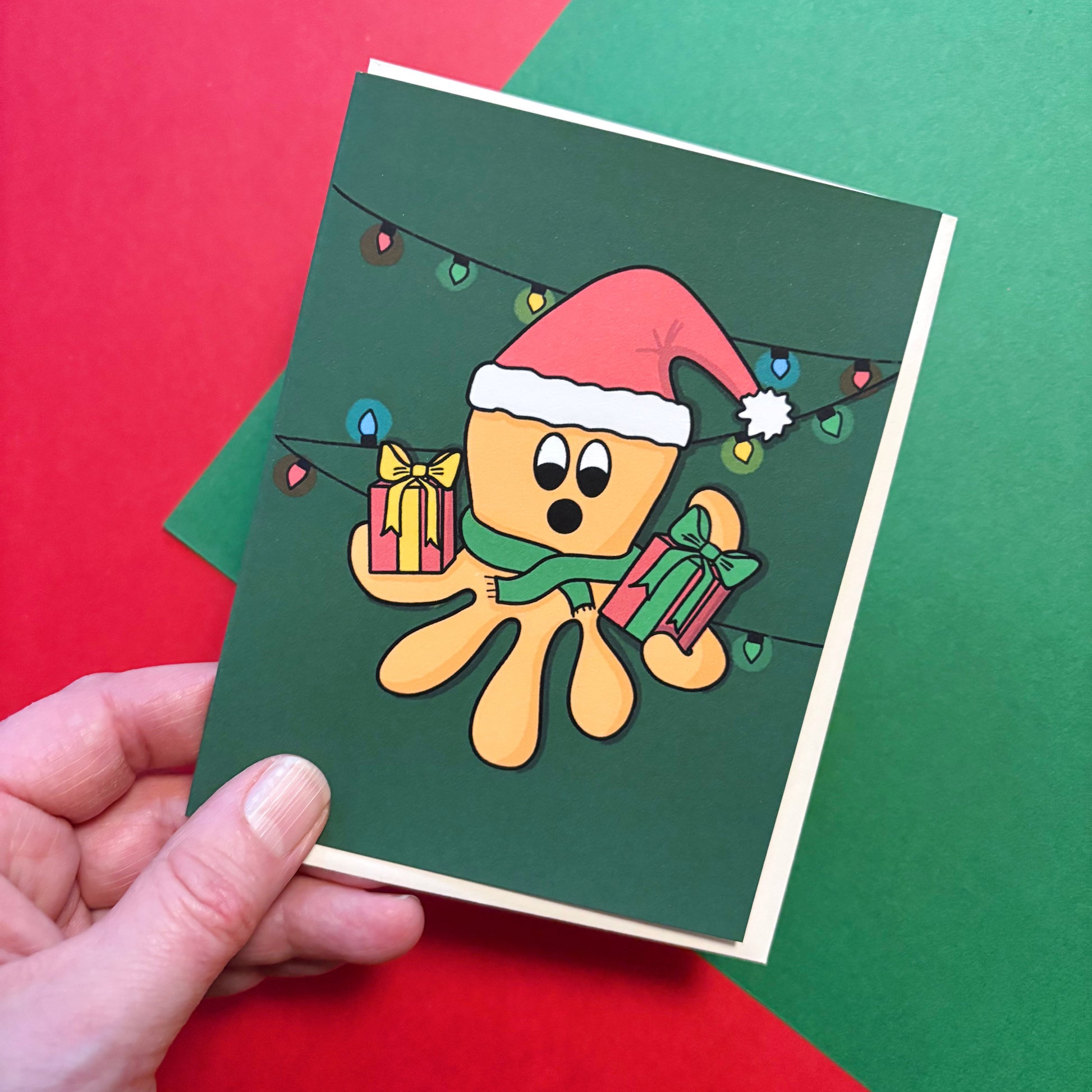 Hand holding a Christmas-themed greeting card with an illustrated orange octopus wearing a santa hat and holding presents in its arms on a red and green background