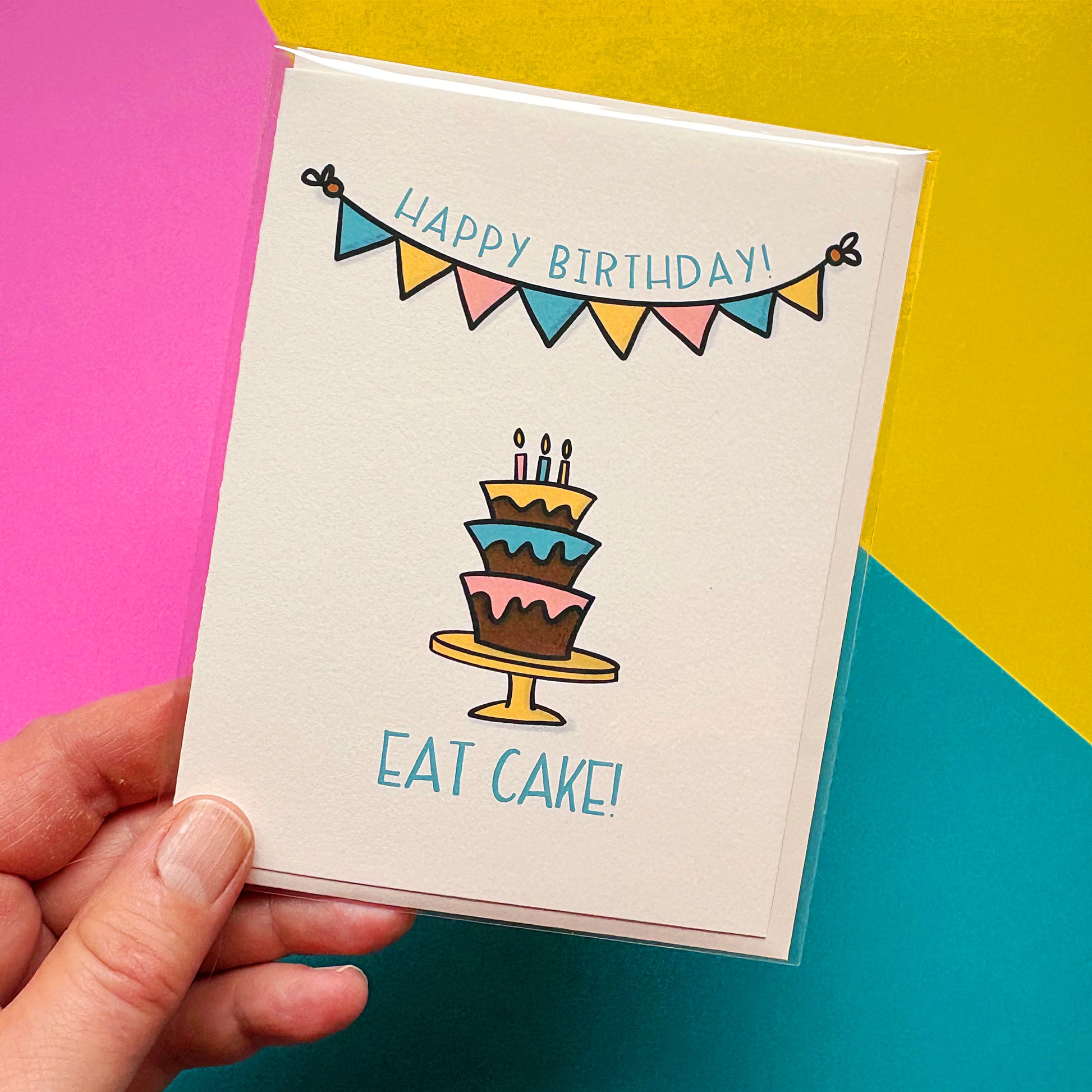 Hand holding a birthday card with a cake illustration and 'Happy Birthday! Eat Cake!' text on a colorful background.
