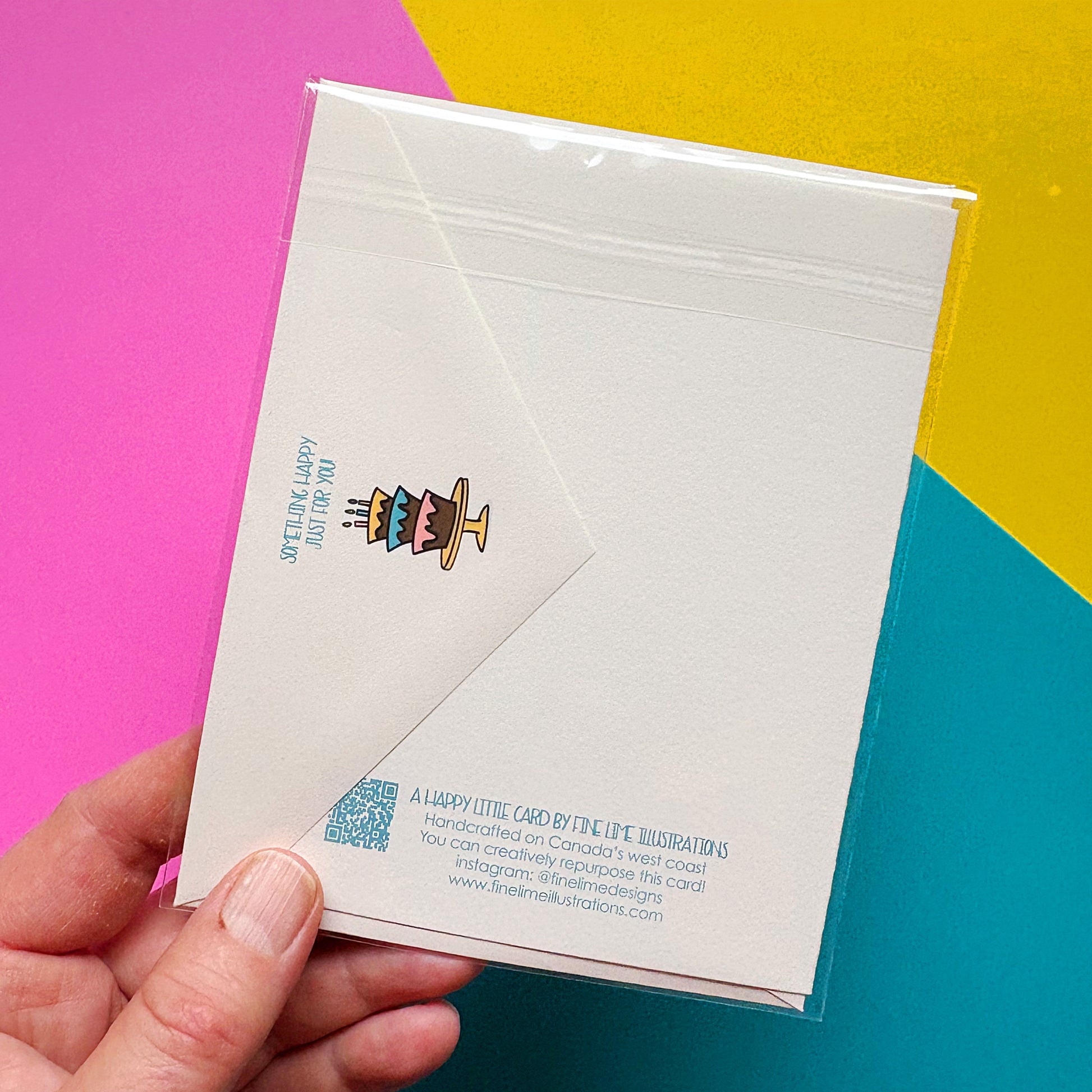 Hand holding a white card flipped to show the envelope flap artwork of an illustrated cake with a colorful design on a multicolored background
