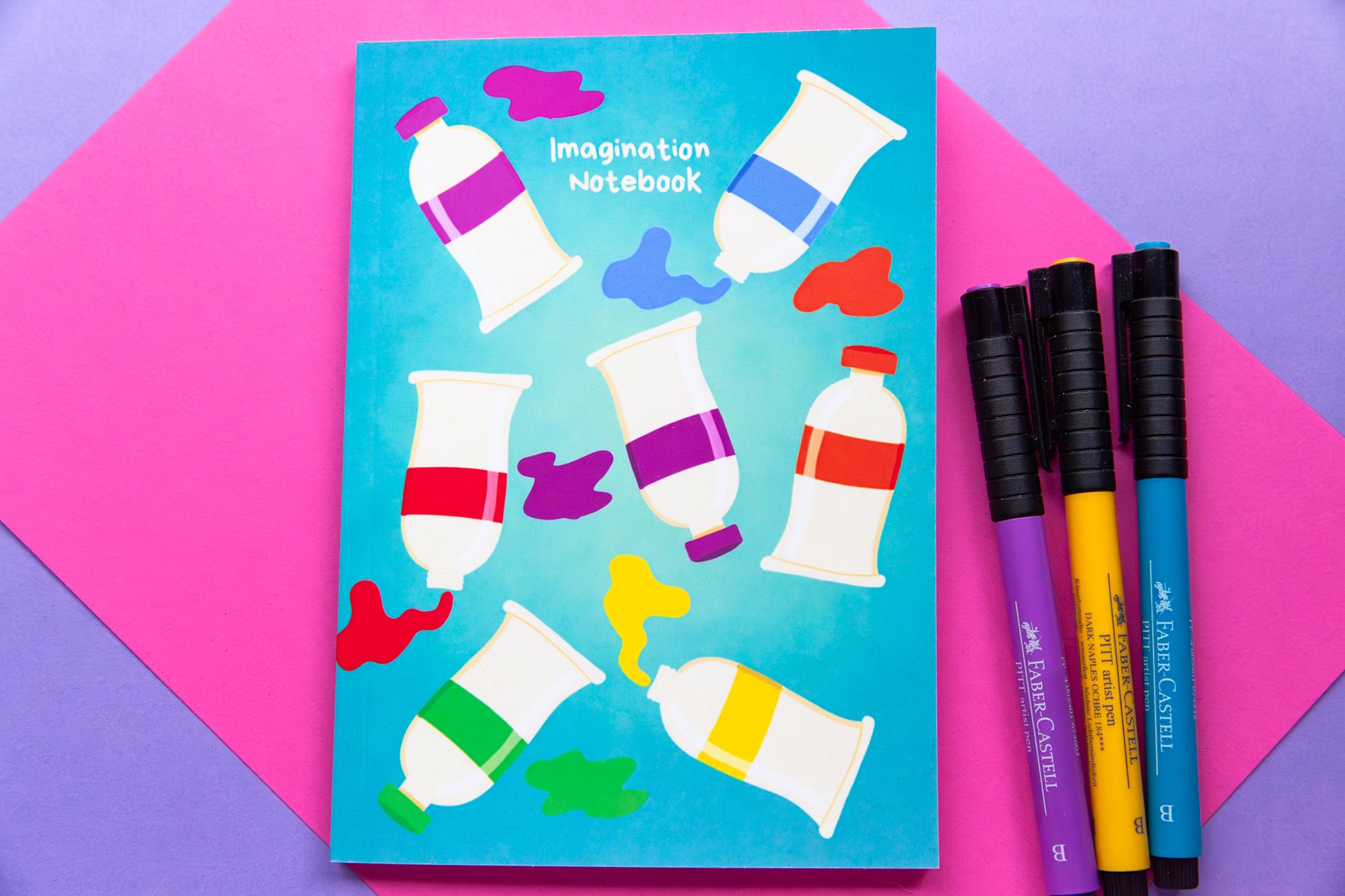 Overhead view of a teal notebook with an illustration of paint tubee and paint splotches in a rainbow of colours. The notebook sits on a colourful pink and purple back drop. Three markers in purple, teal  and  yellow sit next to the notebook