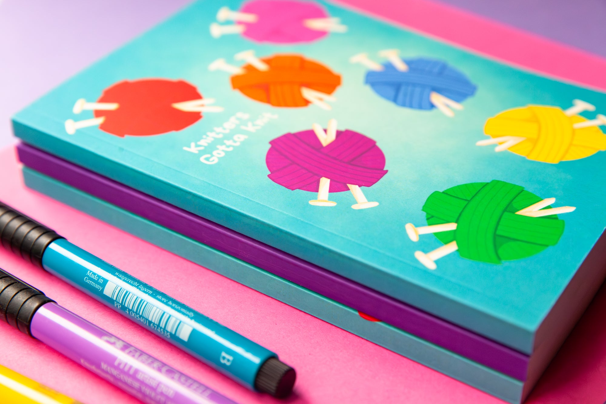 A stack of teal and purple notebooks that shows the thickness of the spine. The top notebook is teal and has an illustration of balls of yarn and knitting needles in an array of colours