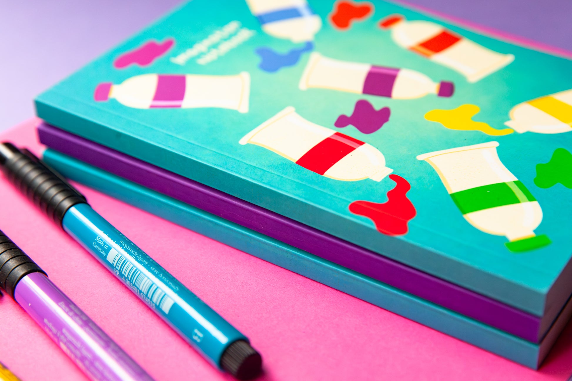 A teal notebook with an illustration of paint tubee and paint splotches in a rainbow of colours on the cover. It's sitting on top of  a stack of notebooks to show the thickness of the spine. The notebook sits on a colourful pink and purple back drop. Three markers in purple, teal  and  yellow sit next to the notebook