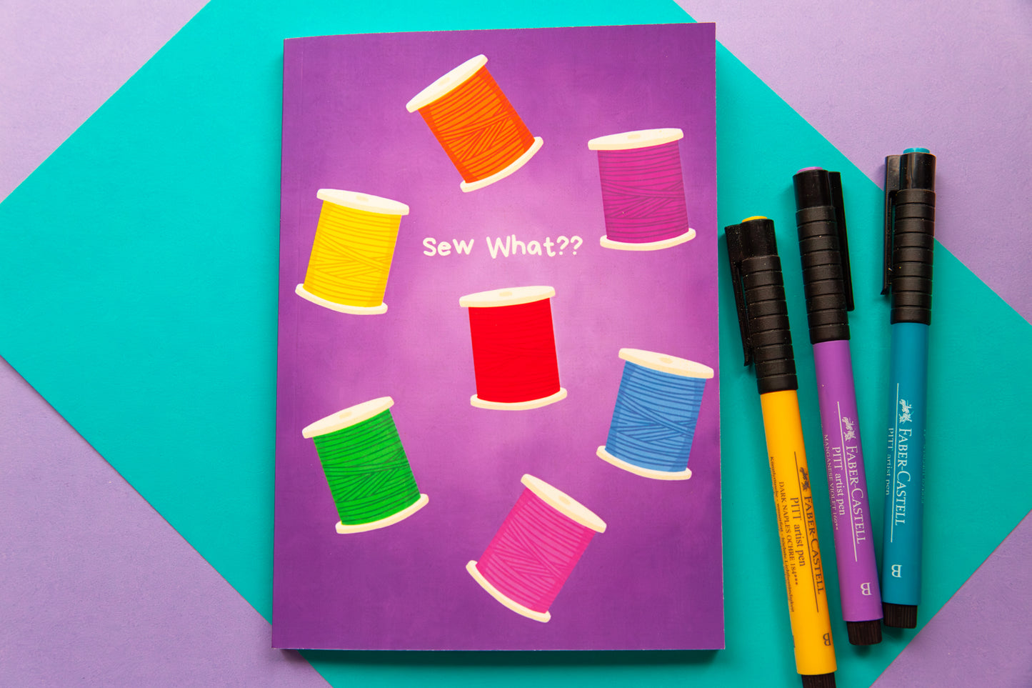 Overhead view of a purple notebook with an illustration of spools of thread in a rainbow of colours on the cover. The notebook sits on a colourful teal and purple back drop. Three markers in purple, teal  and  yellow sit next to the notebook
