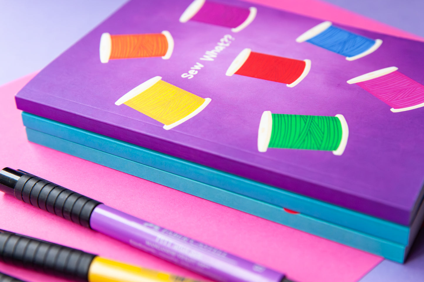 a stack of purple and teal notebooks that shows the thickness of the spine. The purple notebook on the top has a cover illustration of colourful spools of thread in a rainbow of colours
