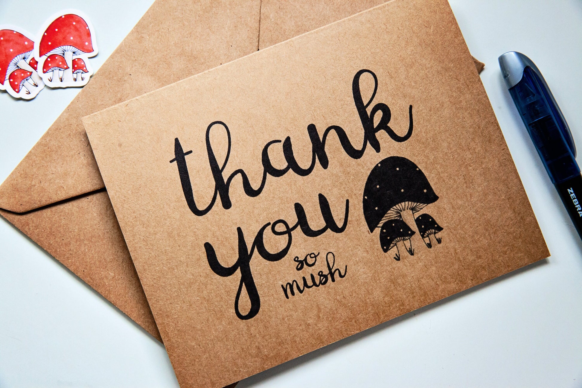 a kraft paper greeting card with matching envelope. The card says "thank you so mush" and has an illustration of a trio of mushrooms.