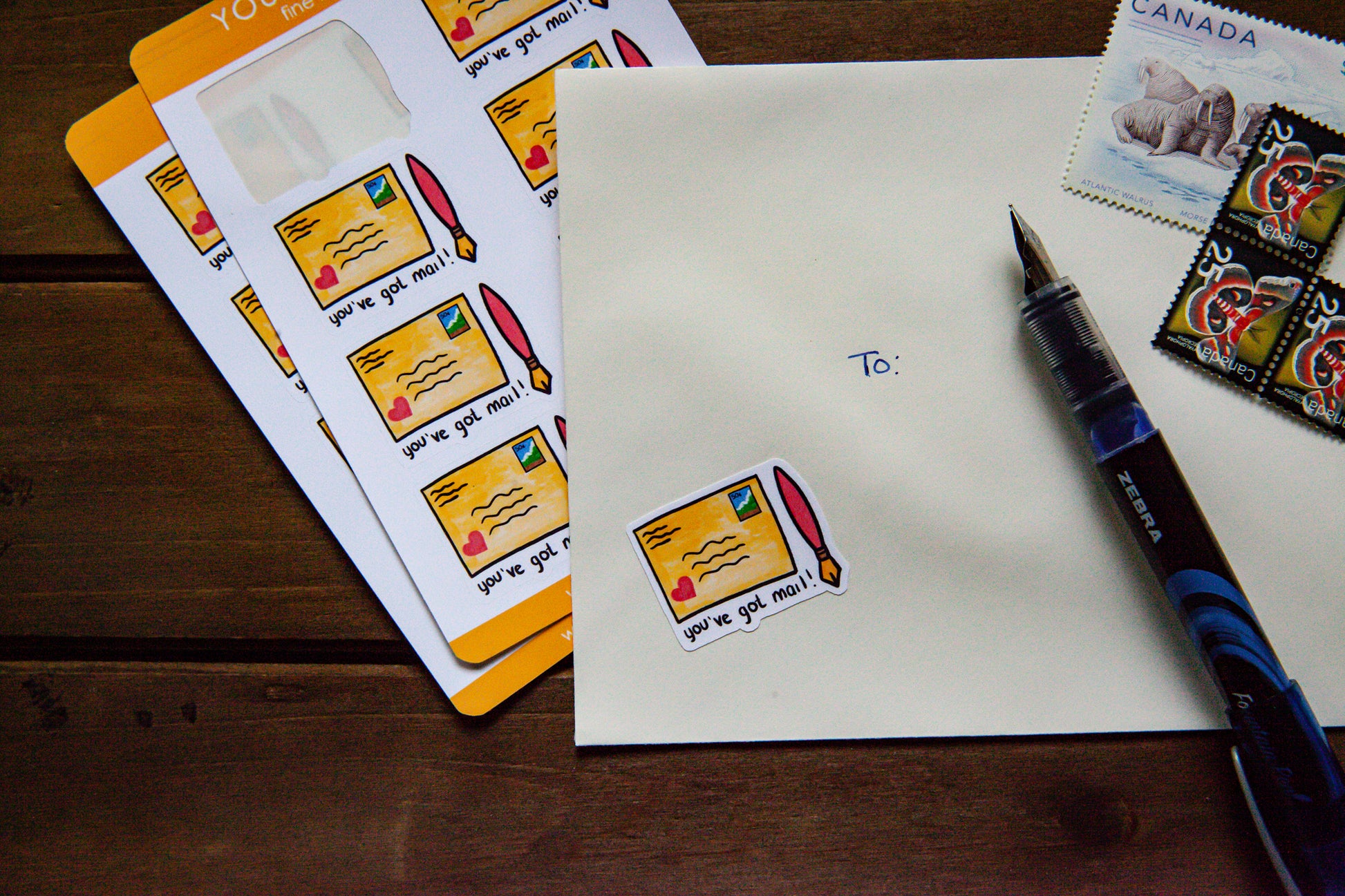 a yellow "you've got mail" sticker on a white envelope with a pen and stamps